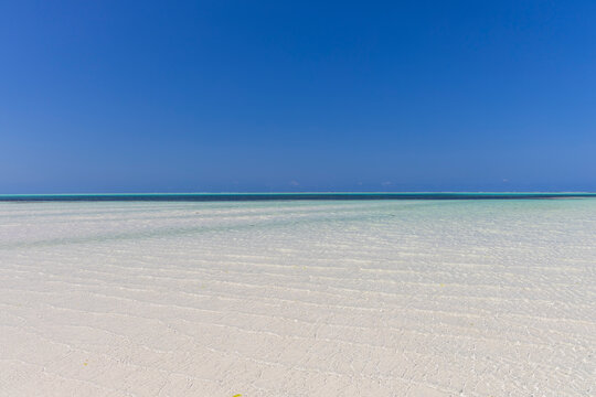 Shallow turquoise water rippling over a vast white sand tidal flat at low tide under a deep blue cloudless sky with the distant reef line on the horizon, Jambiani, Tanzania