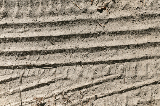 Vehicle tire tracks on sandy ground. Parallel wheel marks in dry soil. Rough earth texture. Transportation trail background. Gray sand surface. Off road terrain. Repeated tread pattern.