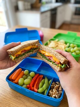 Two hands holding healthy sandwiches with cheese, lettuce, and tomatoes over colorful lunch boxes with snacks
