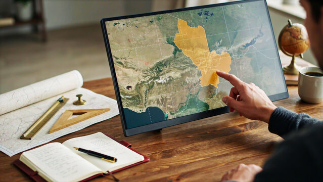 A photograph of a professional cartographer using a digital display and traditional drafting tools to analyze a topographic satellite map on a desk