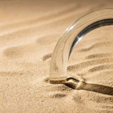 Glass arch shape pressing into textured beach sand with transparent sculpture abstract refraction creative background aesthetic minimalist material contrast clearance structure