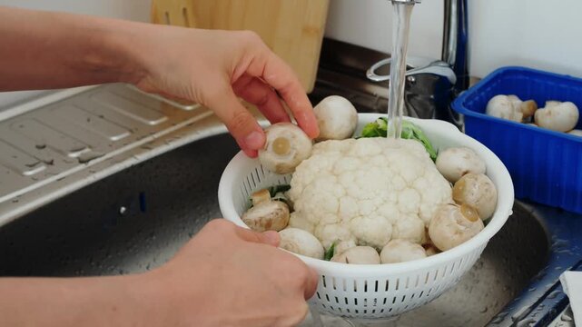 Hands rinse raw vegetables in white colander beneath running water tap as part of healthy home cooking routine. Cook carefully cleans cauliflower head alongside button mushrooms over sink.