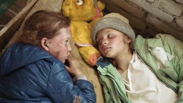 Adolescents lying close on mattress with worn stuffed toy between them, gazing silently with weary eyes, faces marked by dirt and exhaustion, torn jackets and rough wall