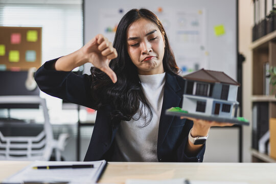Woman agent holding a house model and giving a thumbs down gesture, expressing dissatisfaction with a property or loan deal.