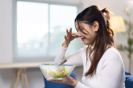 Young woman pinching nose avoiding bad smell. Young Asian woman holding a salad bowl, pinching her nose, avoiding an unpleasant smell or healthy food she dislikes.