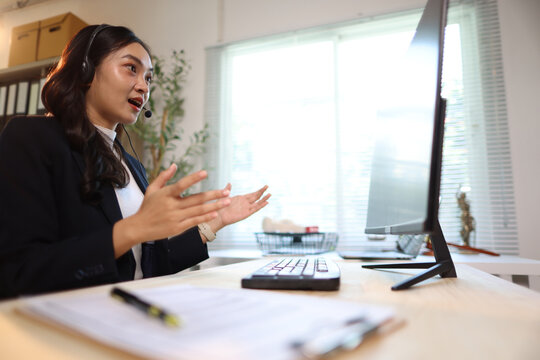 Smiling woman providing customer service work from home. Young Asian woman working from home, wearing a headset, typing on a computer, and smiling while helping a customer.