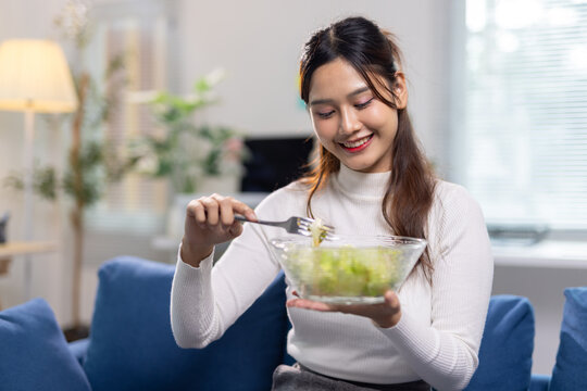 Asian woman enjoying a fresh salad on her sofa, promoting healthy eating, wellbeing, and a balanced lifestyle indoors.