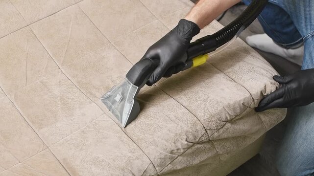 Worker cleans beige sofa cushion seam with extractor nozzle to remove dirt from upholstery fabric.
