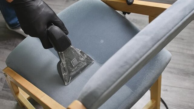 Worker deep cleans gray chair upholstery with extractor nozzle to remove dirt and refresh fabric.