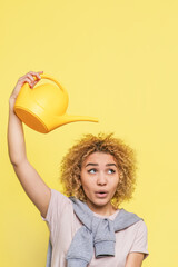 Vertical picture of funny girl. She holds a watering pot upon her hand. She is ready to pour some water on her hair. Girl is looking to the side. Isolated on yellow background. © estradaanton