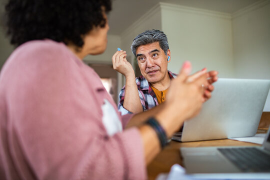 Man discusses with woman at home table, holding an earbud