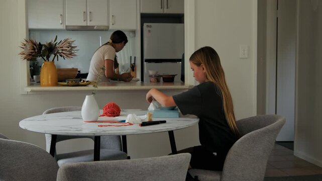 Daughter focuses on her creative art project at the marble table while a mother works in the background of a domestic home space