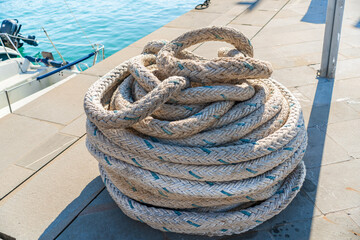 Fototapeta premium A large coil of weathered braided marine rope resting on a harbor quay beside turquoise water and a small boat