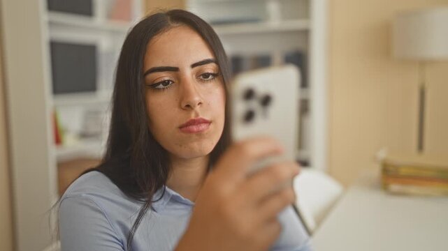Woman holding smartphone and pen to nose while gazing at a screen across a desk in an office with bookshelf and lamp; contemplation pondering reflection.