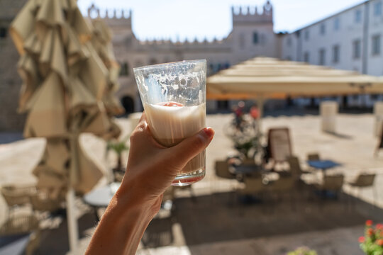 A hand holds a glass of creamy coffee in a cafe overlooking Tito Square and the historic Praetorian Palace in Koper, Slovenia