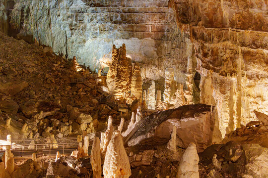 Warm light bathes ancient cave formations. Stalactites hang low, proud and tall. Stalagmites rise from earth, seeking sky. Rock walls glow with earthy hues and textures. Quiet wonder fills the dim.