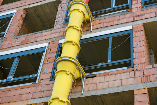 Yellow construction debris chute attached to an unfinished brick building with exposed concrete floors, window frames, chains, and metal supports