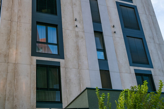 Building facade with beige stone panels, dark blue framing, tall windows, horizontal blinds, exterior lights, and green foliage below