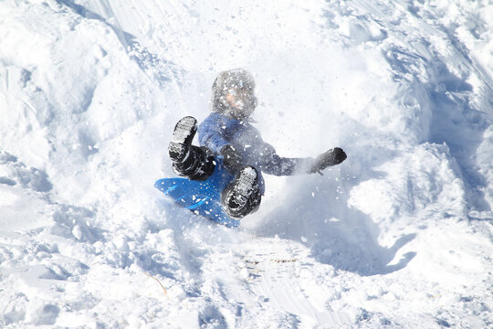 A thrilling action shot capturing a young boy sledding down a steep, snow-covered hill on a bright blue plastic sled. Surrounded by kicked-up snow powder and bright winter light, the dynamic scene hig