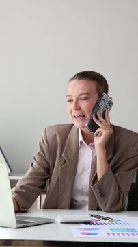 Businesswoman talking on smartphone while using laptop computer at her desk in a modern corporate office workplace