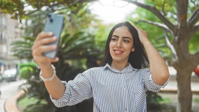 Woman holds smartphone at arms length while smiling and touching her hair on a city street lined with trees; confidence connection.