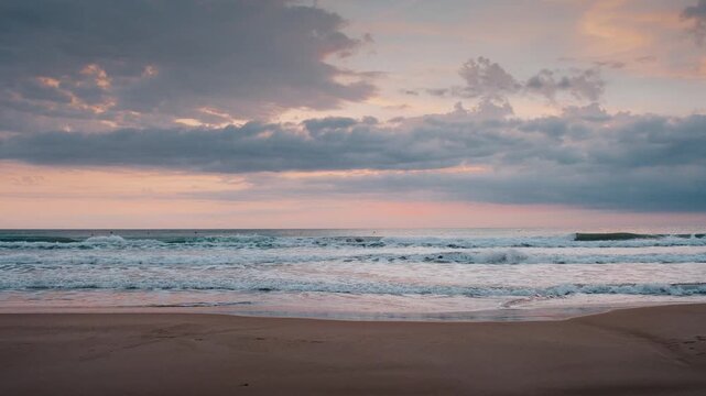 Beautiful sunrise over gentle sea waves rolling onto a sandy beach under soft pastel clouds during dawn.