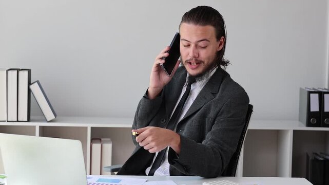 Young businessman with long hair talking on the phone and playing with a pen while sitting at his office desk