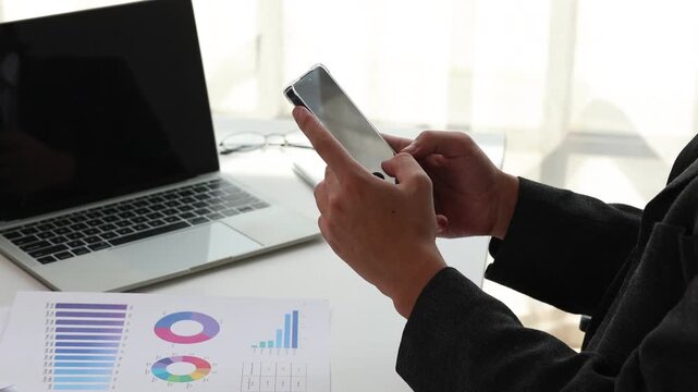 Businessman using a smartphone at his desk with laptop and financial documents for business analysis and marketing strategy