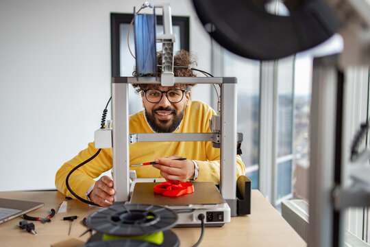 A smiling Indian man with glasses performs routine maintenance on a 3D printer, checking its components and preparing it for use