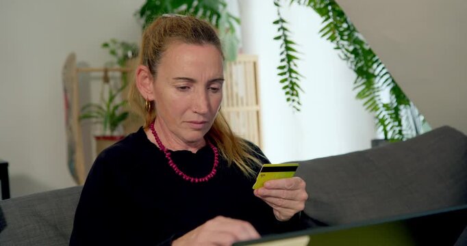 Focused Woman Shopping Online: A woman intently uses her credit card and laptop at home, suggesting online purchasing or banking.