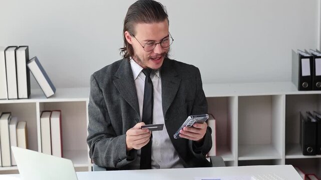Excited businessman making an online payment with a credit card and smartphone while celebrating success in the office