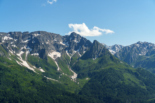 Majestic mountain range rises under clear blue sky. Snow patches cling to rocky peaks. Green forests blanket lower slopes. Single white cloud drifts lazily. Ideal for travel, nature.
