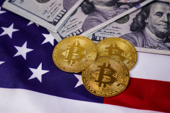 Three physical golden Bitcoin token coins arranged over the fabric of the US national flag, with a spread of 100 dollar in the background.