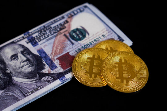 Two physical golden Bitcoin token coins arranged overlapping horizontally next to a thick stack of United States 100 dollar banknotes.