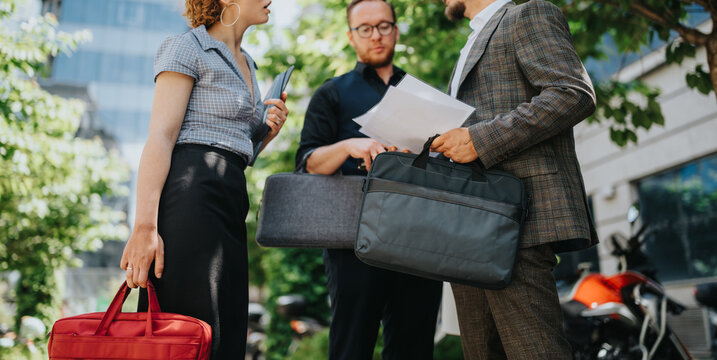 Three business colleagues stand outside a modern office building holding documents and briefcases. The group discusses papers during an outdoor work meeting in a city setting.