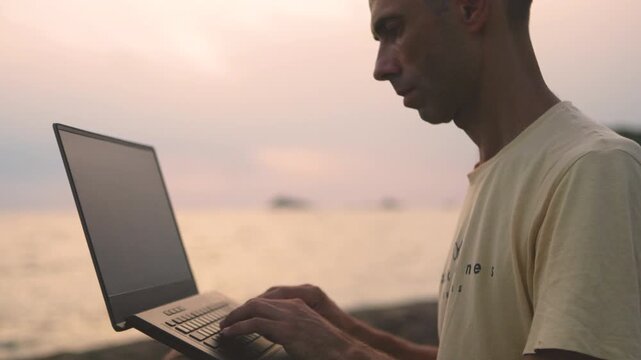 Man with laptop on pebble shoreline overlooking calm ocean at dusk