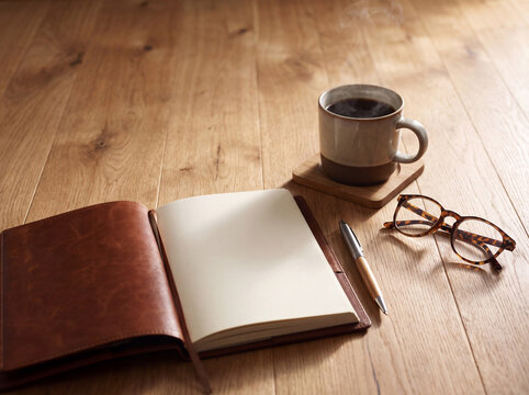 A cozy workspace setup with an open notebook, steaming coffee, stylish glasses, and a pen on a warm wooden table.