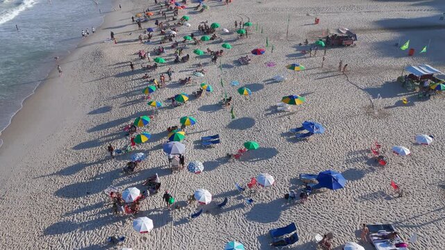 Aerial flyover of Copacabana Beach with beachgoers and colorful umbrellas dotting the sand on a sunny day, Rio de Janeiro, Brazil.