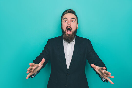Berded man is absolutely amazed. He is looking to camera awith his mouth opened. Also he is showing his hands. Isolated on blue background.