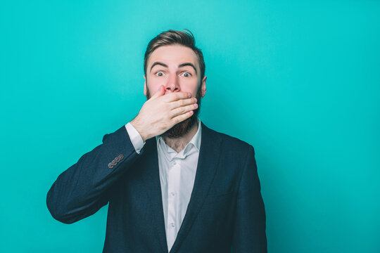 Bearded guy is closing his mouth with the hand. He looks scared and terrified. Isolated on blue background.