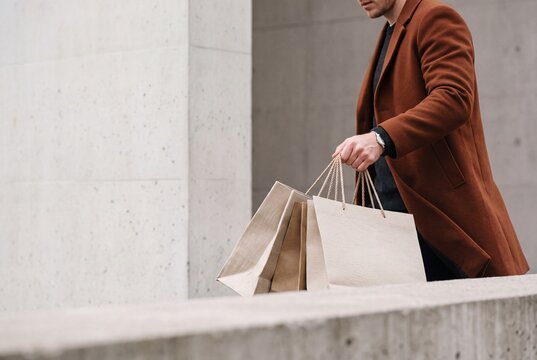 Stylish well dressed man wearing an elegant coat carries paper shopping bags while walking near modern architecture outdoors