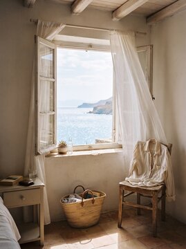 Coastal summer room with an open window, sheer curtains, blue ocean view, linen shirt, wooden chair and woven bag, captured in a warm candid travel diary photography style.