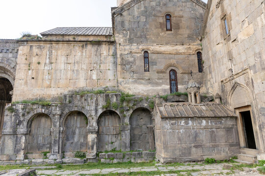 Weathered stone courtyard wall of Tatev Monastery with four blind arches and a carved tomb chapel beside tall arched windows and a doorway with weeds, Tatev, Armenia