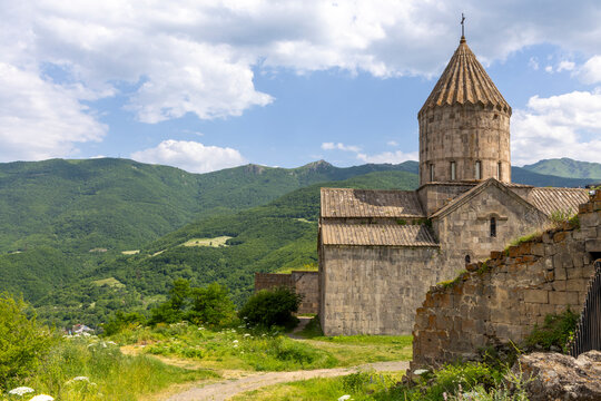 Tatev Monastery church with a cylindrical drum and fluted conical dome standing above forested green mountains and a dirt path under a bright cloudy blue sky, Tatev, Armenia