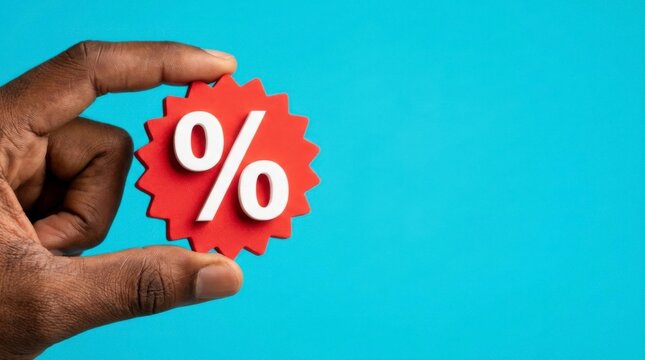 Dark-skinned hand holding a red percentage sign with white symbol against a vibrant blue background, representing discounts or interest rates