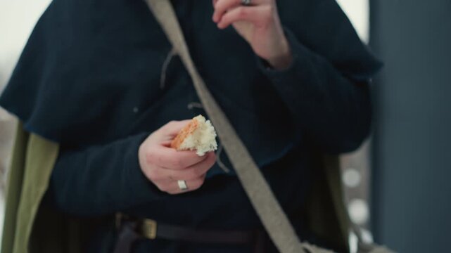 Person wearing medieval style dark blue garments and rings on their hands eats piece of bread. subject holds partially eaten item breaks off another small portion by hand and brings