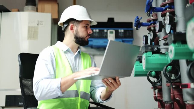 Diligent bearded engineer, wearing a white hard hat and reflective vest, intently inputs data on a laptop, examining complex boiler room equipment and pipes.