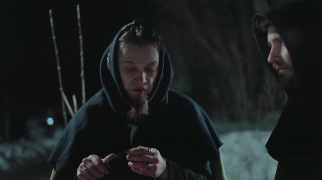 Two people dressed in heavy cloaks and hoods stand outdoors at night examining something held between their hands. setting is dark with snow covered ground visible suggesting cold