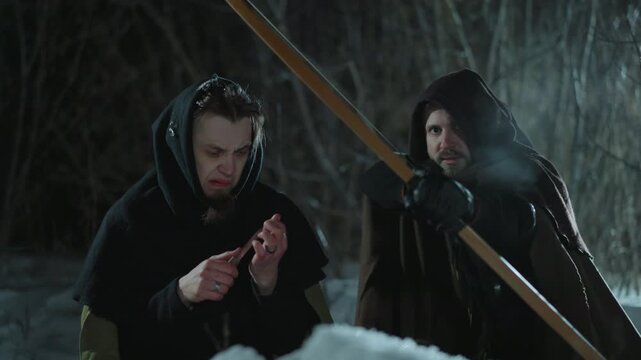 Two men in hooded cloaks one examining knife and other holding arrow stand together at night with dark forest background wolf hunting fantasy werewolf