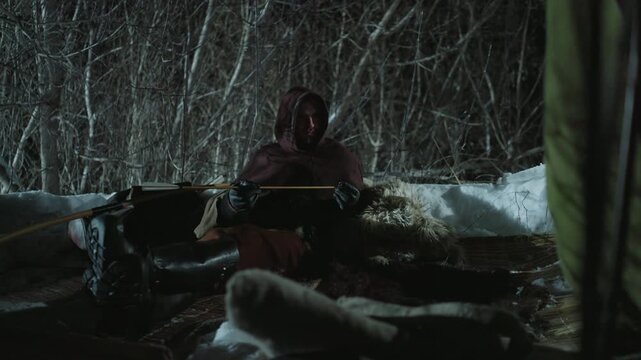Two people in hooded cloaks sit inside makeshift snow covered shelter at night one holding arrow and other resting beside what appears to be prepared prey. scene is set against dense